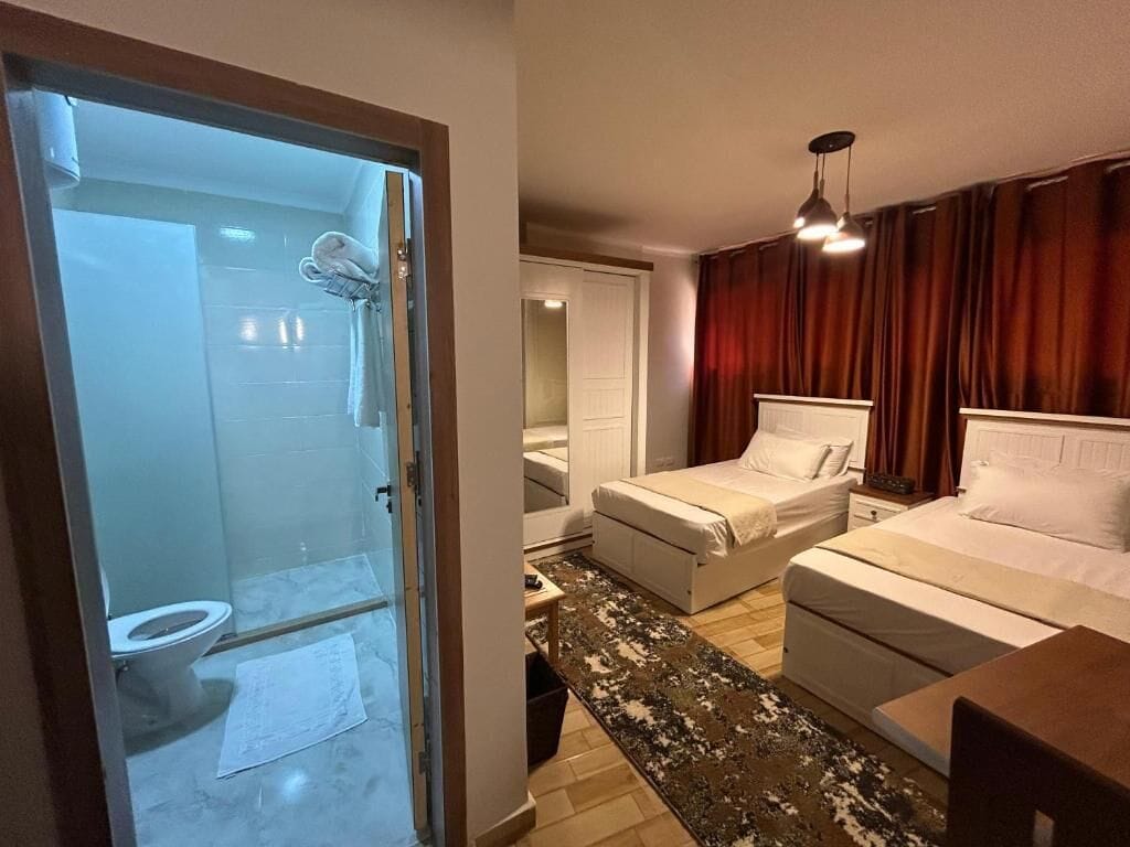 A hotel room with a glass-enclosed shower, a bed with a white headboard, a bedside table, and a small table with a chair.