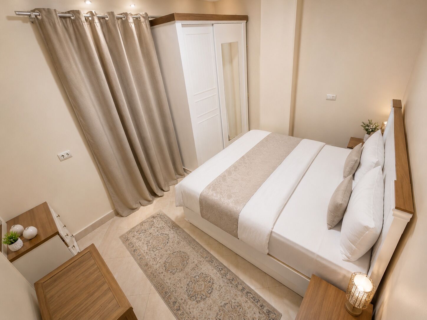 A bedroom with a bed, a nightstand, a bedside table with a lamp, a wooden dresser, a rug, and a door with curtains.
