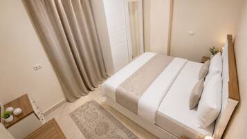 A bedroom with a bed, a nightstand, a bedside table with a lamp, a wooden dresser, a rug, and a door with curtains.