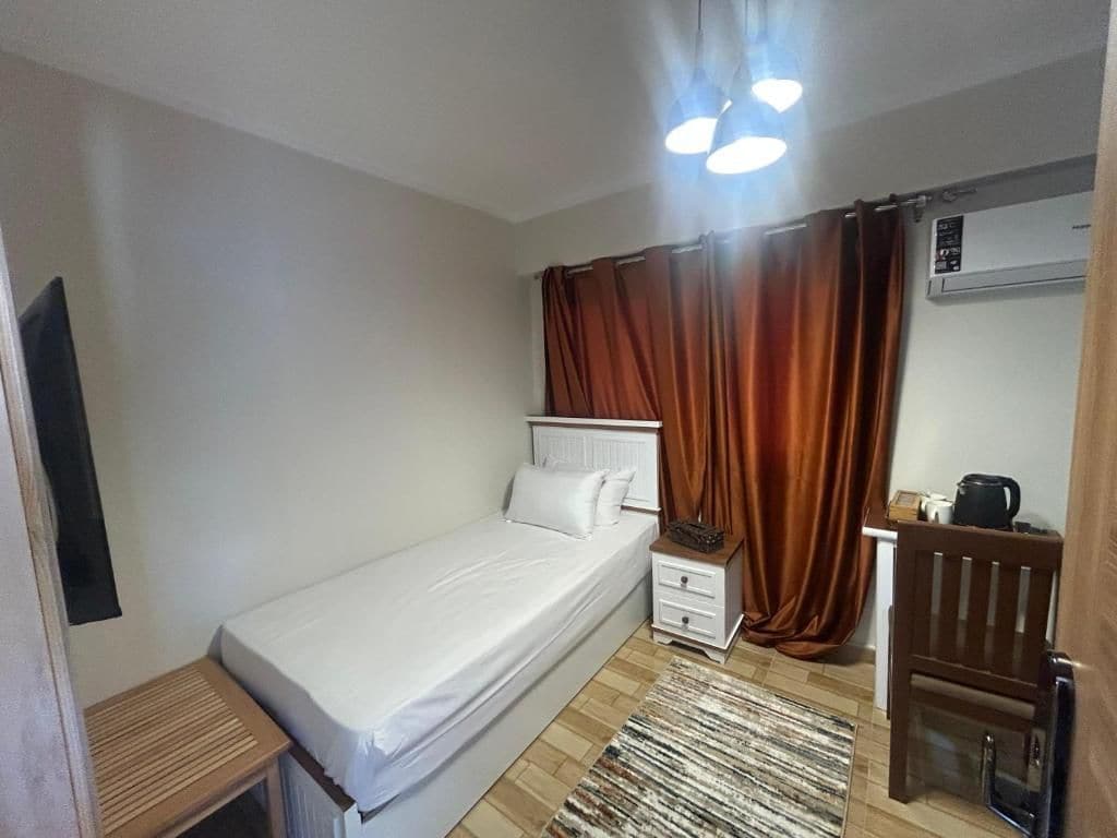 A single bed with a white mattress and pillows, a small bedside table with drawers, a brown curtain, a wall-mounted air conditioning unit, and a small cabinet with a kettle.