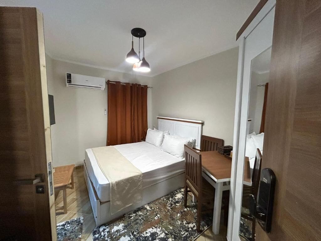 A bedroom with a bed, a desk, a chair, an air conditioning unit, and a mirror.