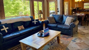 A cozy living room with a sofa and a coffee table, large windows with a view of trees, and a wooden dining area. - Log Cabin w/ Billiards & Table Tennis - Mtn Views, 24mi Breck, Pet-Friendly (Fairplay)