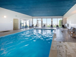 An indoor swimming pool with clear water, surrounded by lounge chairs and a view of the outdoors.