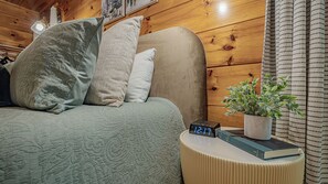A cozy room with a bed, pillows, a bedside table with a clock, a book, and a potted plant.