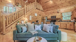 A cozy cabin interior with a wooden staircase, a sofa with cushions, a kitchen area with wooden cabinets, and a dining table.