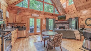 A cabin interior with a kitchen, dining area, and living room, featuring wooden walls, a stone fireplace, and large windows.