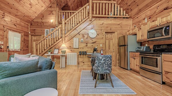 A log cabin interior with a staircase, wooden walls, a kitchen area, and a seating area with a patterned chair.