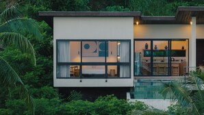A modern house with large windows, a visible kitchen area, and lush greenery surrounding it. - DUNIA LOMBOK (Selong Belanak)