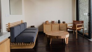 A modern living room with a dark wood floor, a blue sofa, a wooden coffee table, a wicker cabinet with decorative items, and a glass door leading to a stone wall. - DUNIA LOMBOK (Selong Belanak)