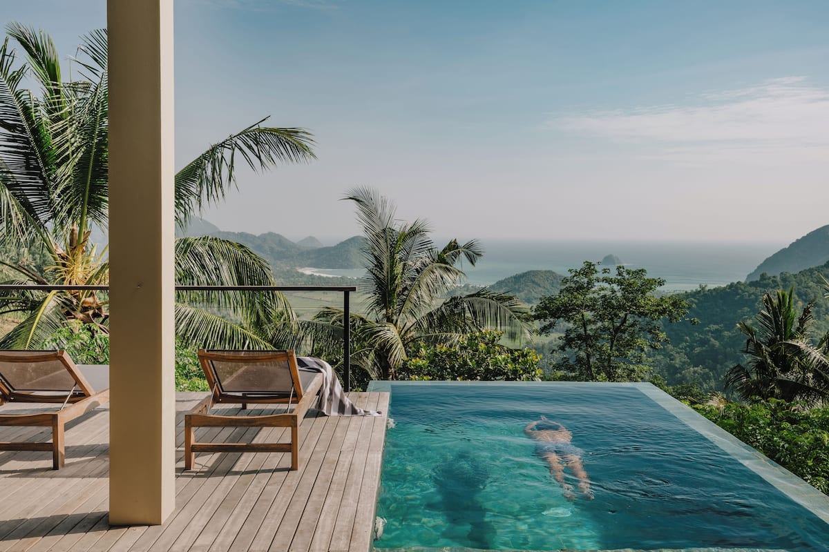 Une piscine offrant une vue dégagée sur l’océan et les montagnes, entourée de palmiers et de chaises longues en bois.