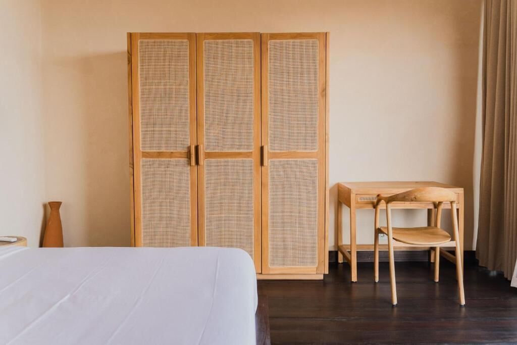 A room with a bed, a wooden wardrobe with a woven front, a wooden chair, and a small table.