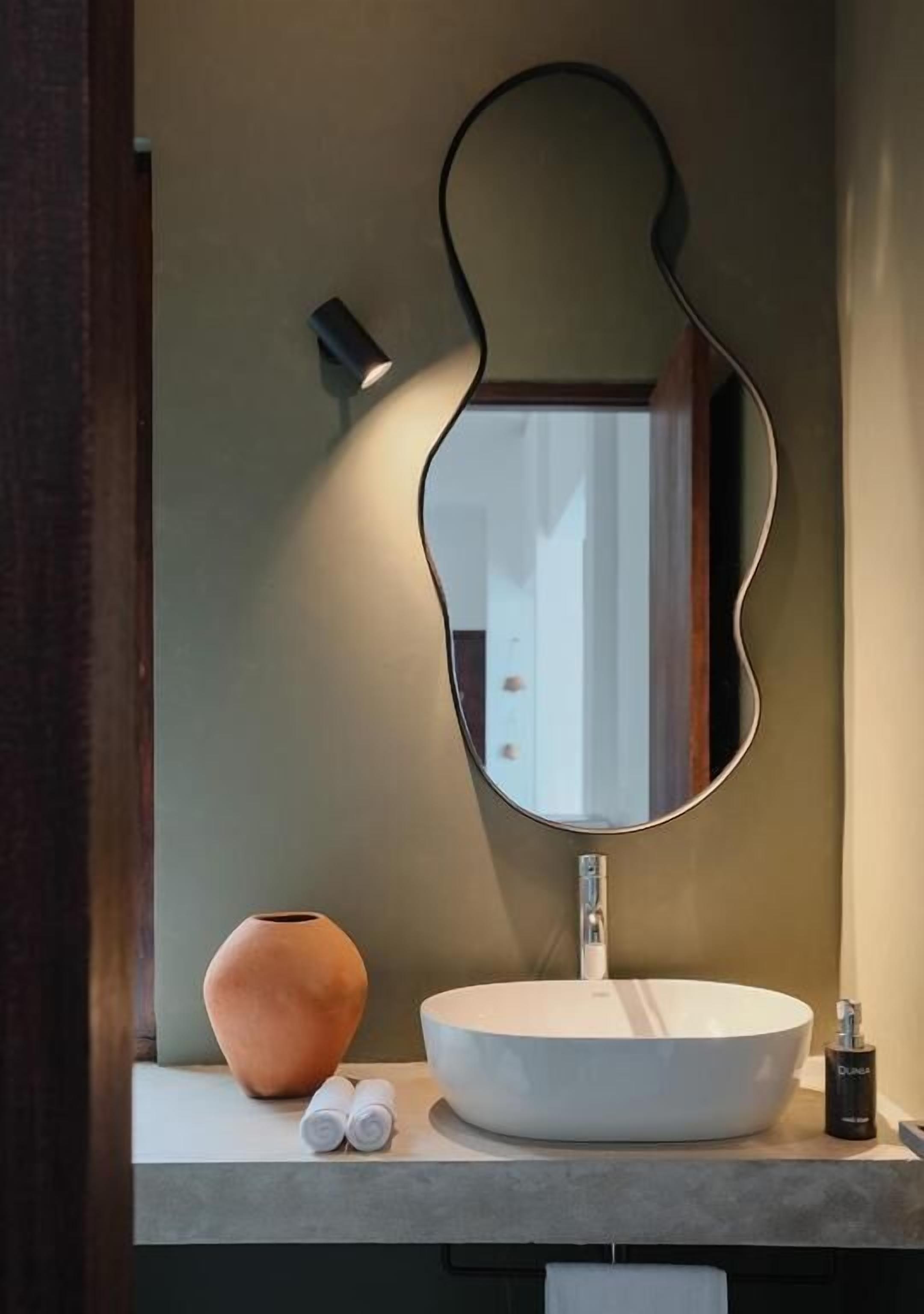 A bathroom vanity with a round white sink, a large mirror with a wavy frame, a small vase, and two rolled towels.