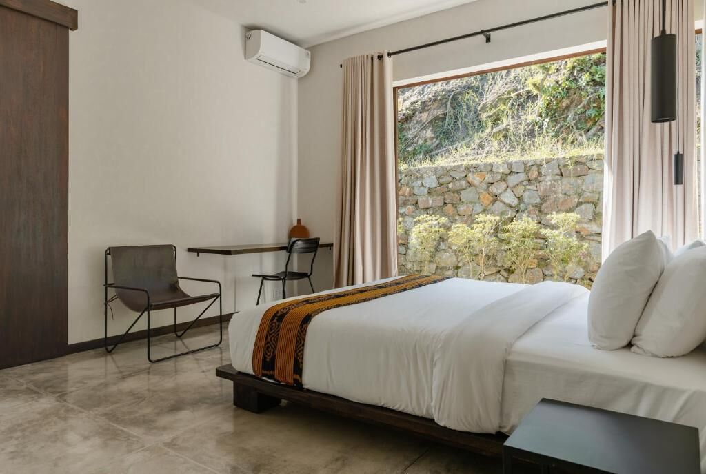 A bedroom with a bed, a desk, and a chair. There is a window with curtains, a stone wall outside, and an air conditioning unit on the wall.