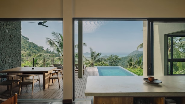 A modern outdoor dining area with a wooden table and chairs, a stone wall, and a view of a lush landscape and mountains. - DUNIA LOMBOK (Selong Belanak)