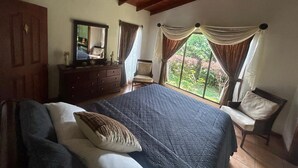Ein Schlafzimmer mit einem großen Bett, einem Schminktisch, einem Sessel und einem Fenster mit Vorhängen.