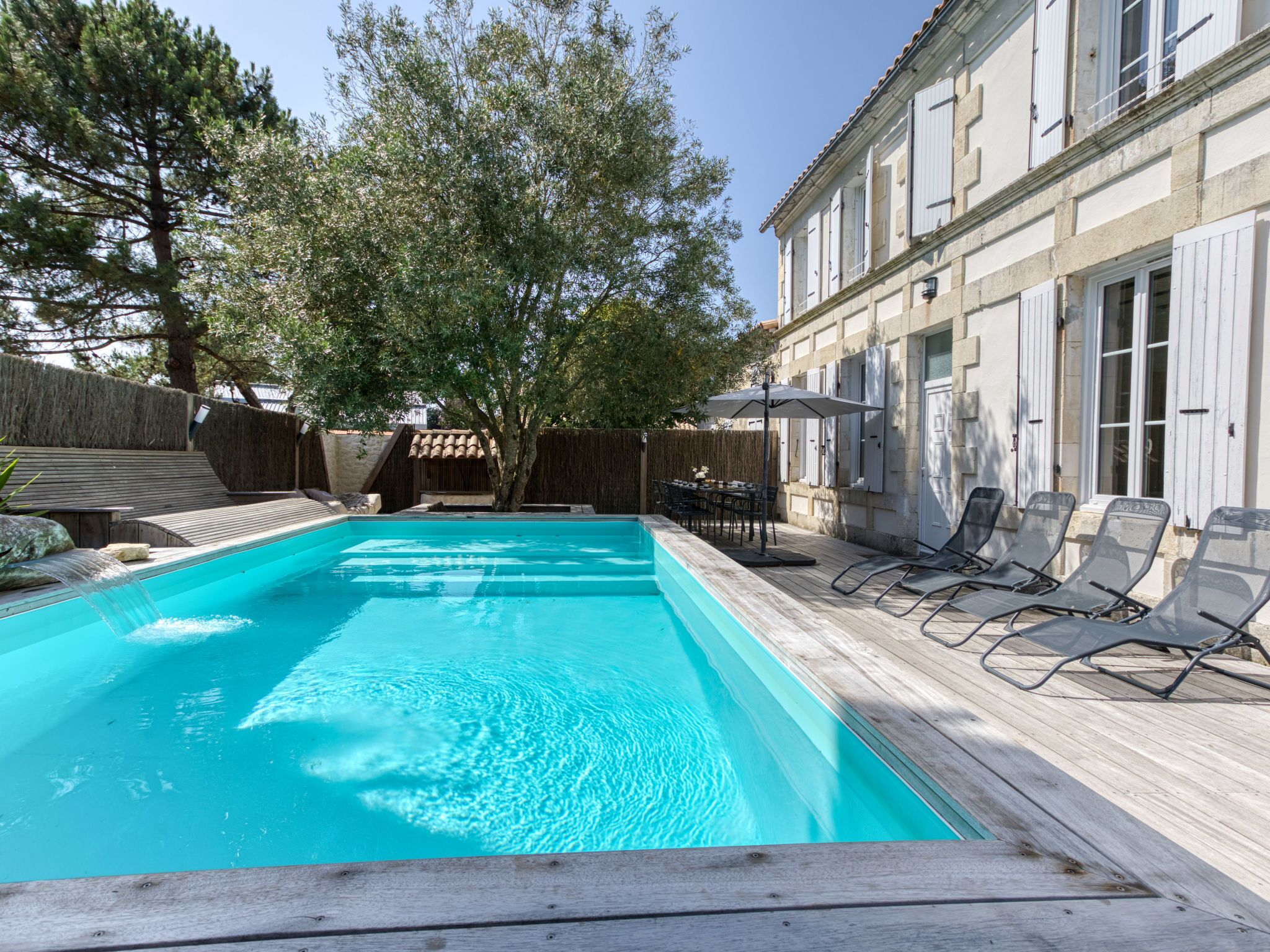 Ein Schwimmbad mit Holzterrasse, Liegestühlen und einem Gebäude mit weißen Fensterläden.
