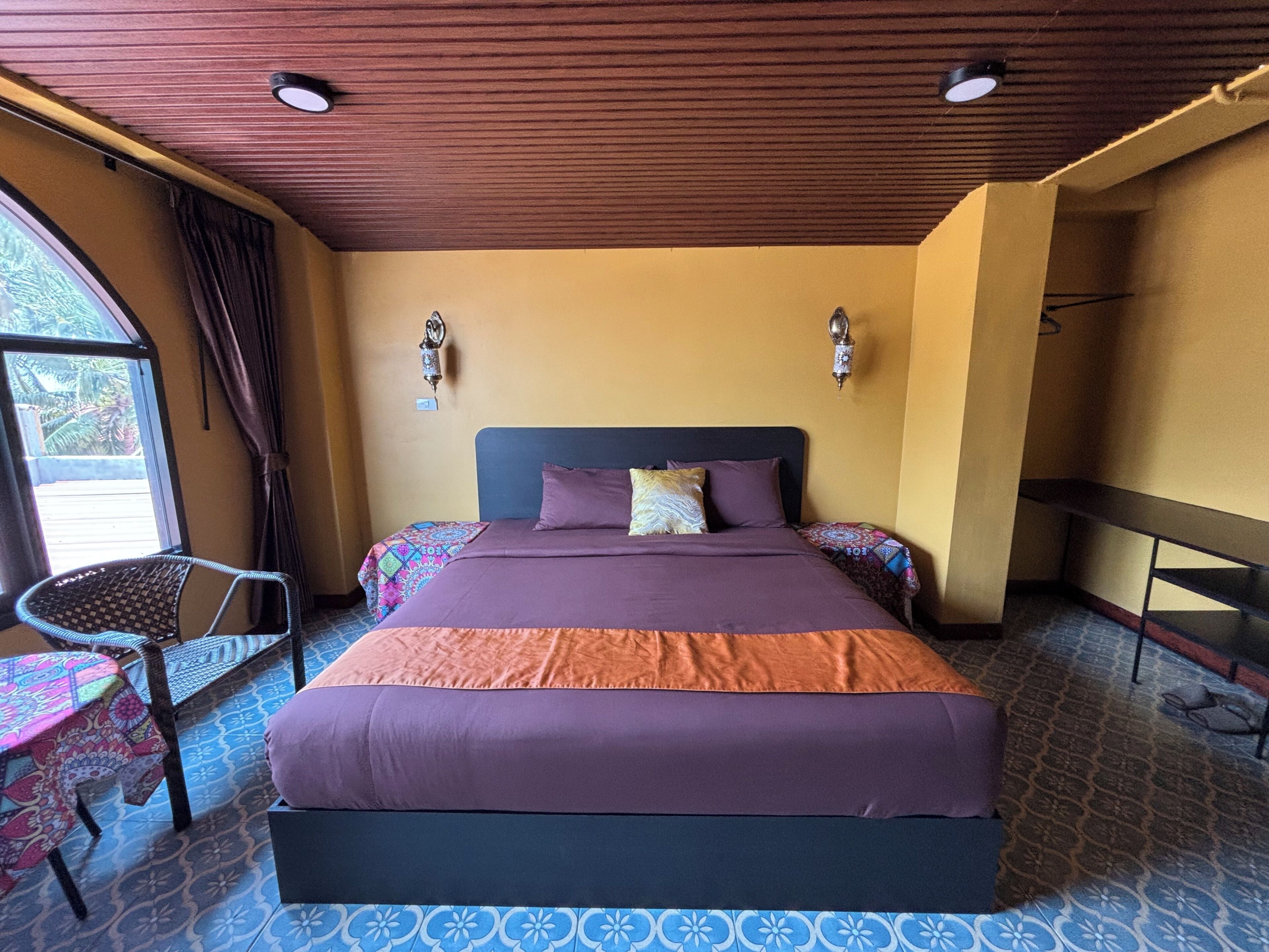 A room with a bed, a chair, a window, and a ceiling with wooden panels.