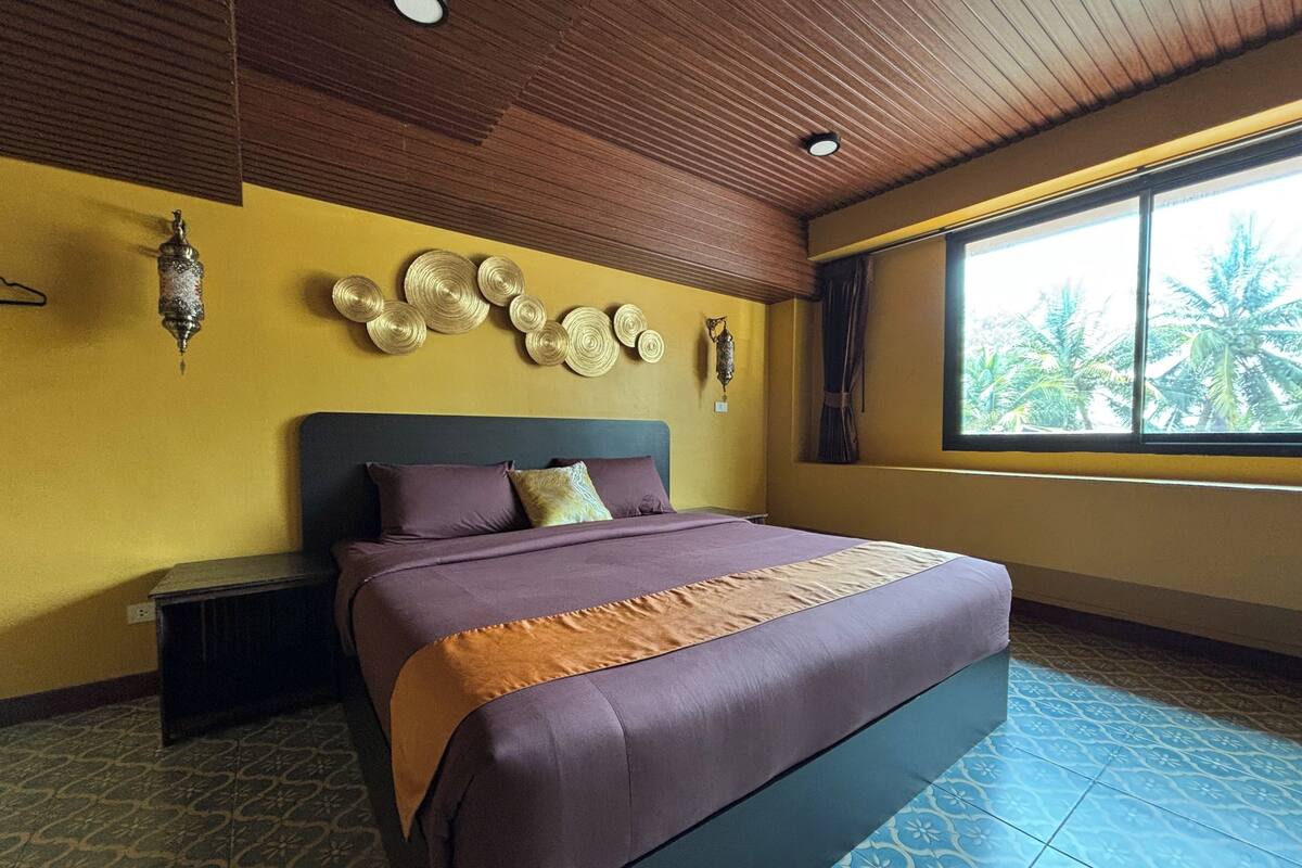 Une chambre avec un lit, une table de chevet, un mur décoratif, une fenêtre donnant sur des palmiers et un sol à motifs.