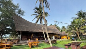 A rustic outdoor seating area with wooden benches and tables, a thatched-roof building, and palm trees. - Phoenix Room (Pathio)
