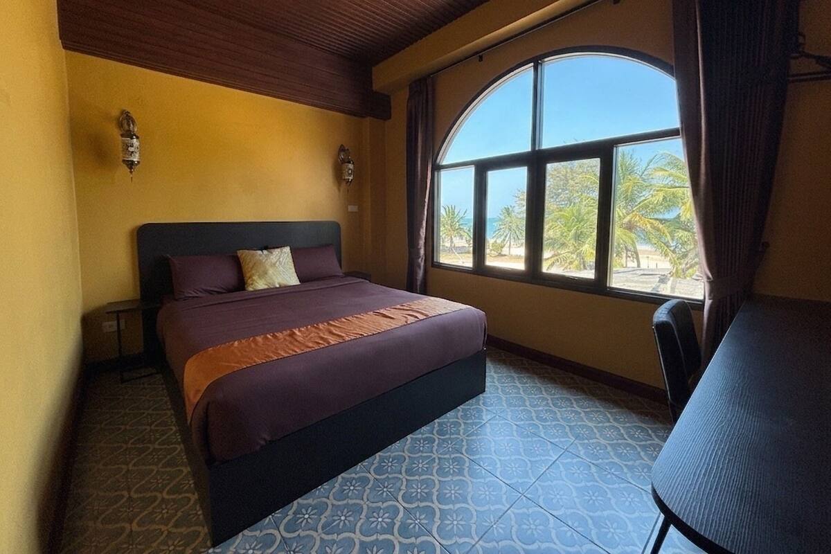 Une chambre d’hôtel avec un lit, une fenêtre cintrée donnant sur des palmiers, une petite table et des carreaux de sol à motifs.