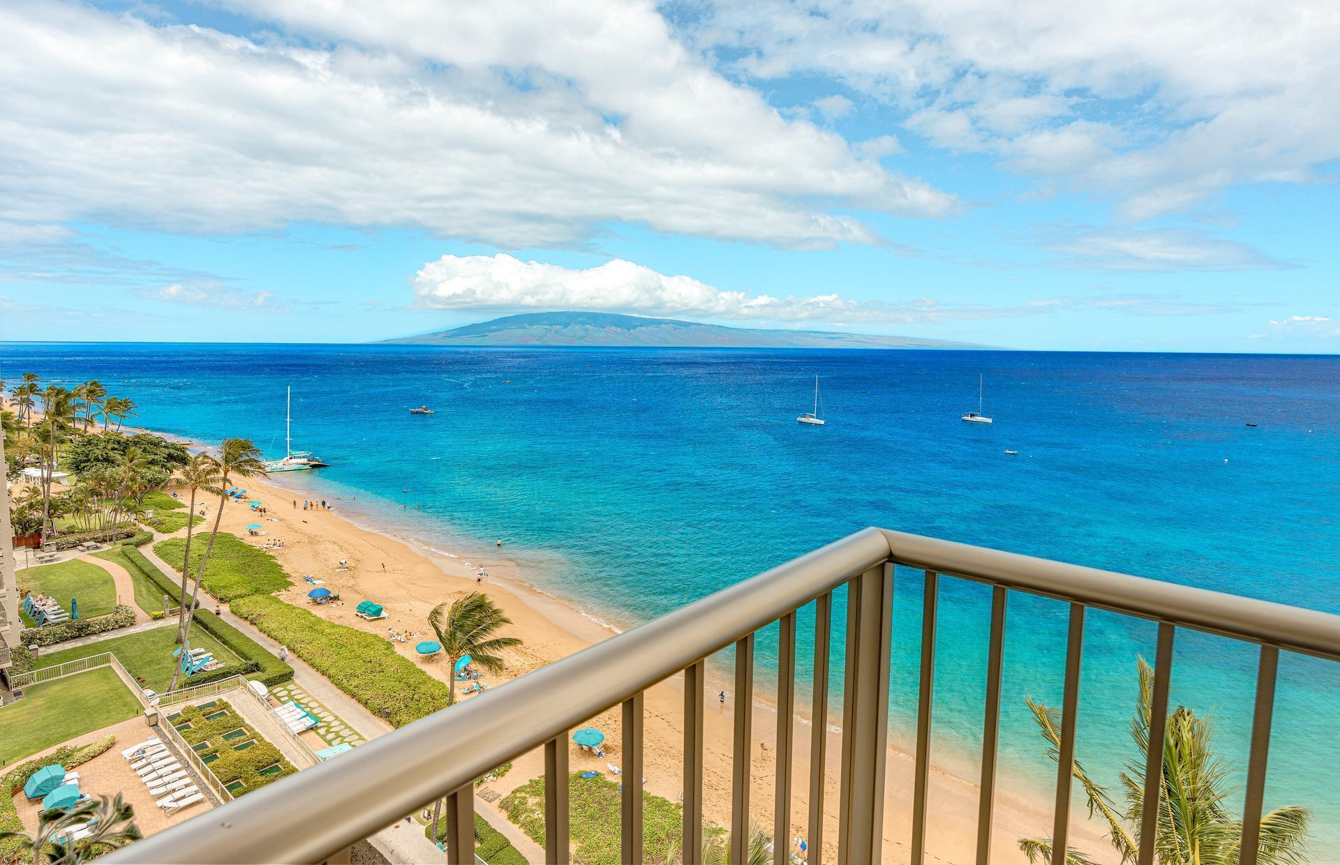 Une vue sur la plage depuis un balcon, avec un ciel dégagé, des palmiers et des bateaux sur l’eau.