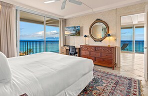 A hotel room with a bed, desk, dresser, and a large window with a view of the ocean.