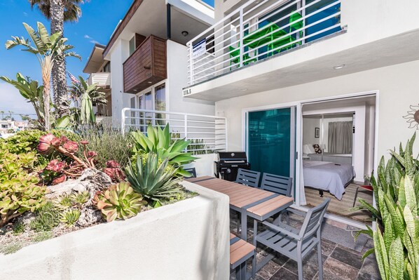 A two-story building with a balcony, a patio with a table and chairs, and a bedroom with a bed and a window.