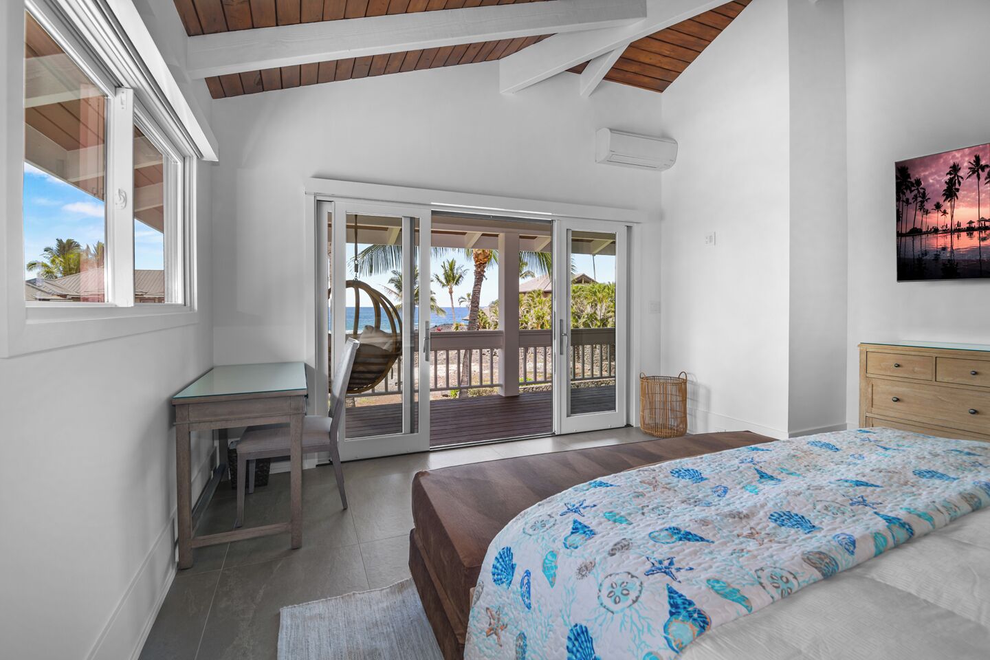 A bedroom with a bed, a desk, and a chair, a balcony with a view of palm trees and the beach, and a painting on the wall.