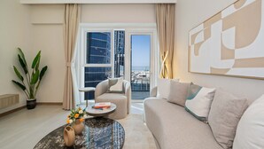 A modern hotel room with a sofa, a round coffee table, a potted plant, and a view of the cityscape through large windows.