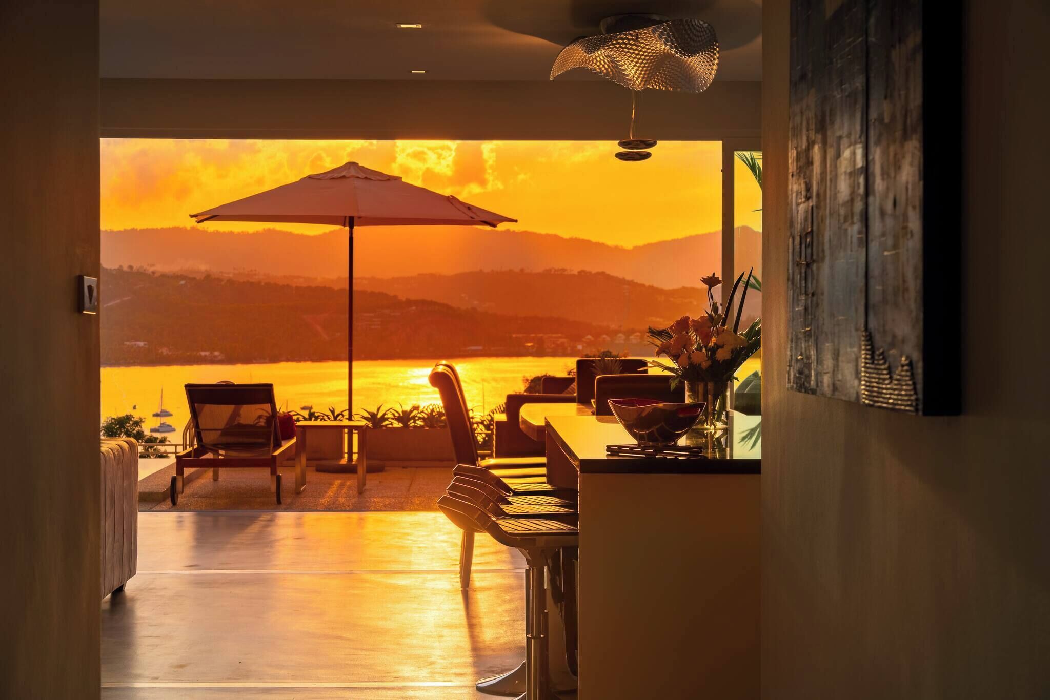 Une pièce avec vue sur un coucher de soleil se reflétant sur une étendue d’eau, un coin repas avec des chaises et une table, et un ventilateur de plafond.