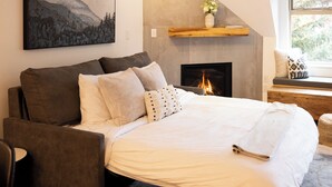 A bedroom with a bed, a fireplace, a mounted painting, and a window with a view of greenery. - Modern Air-Conditioned Ski In/Out 1 BR: Sleeps 4, King bed, Hot Tub, Pool! (Whistler)