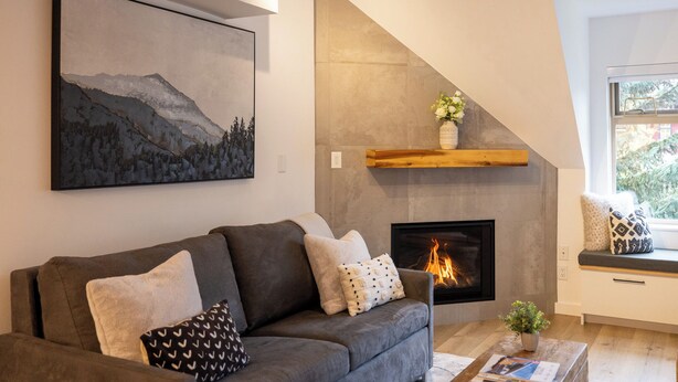 A modern living room with a gray sofa, a fireplace, a wooden shelf, and a large painting of mountains. - Modern Air-Conditioned Ski In/Out 1 BR: Sleeps 4, King bed, Hot Tub, Pool! (Whistler)