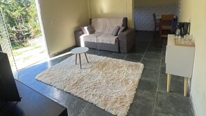Ein Wohnzimmer mit einer Couch, einem Couchtisch und einem Teppich. Eine Tür führt auf einen Balkon mit Blick auf Grünflächen.