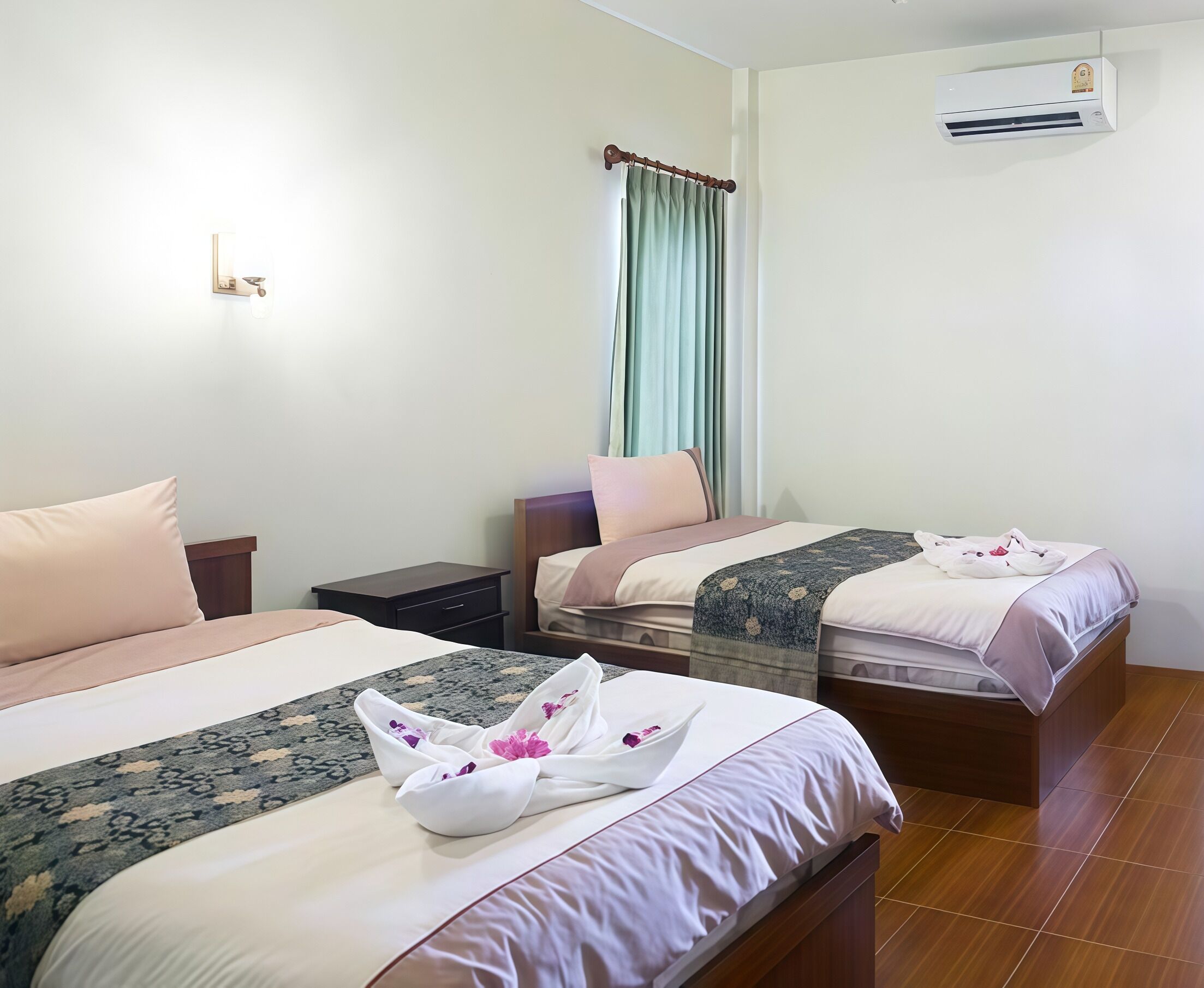 A hotel room with two beds, a nightstand, a small table, an air conditioning unit, and a window with curtains.