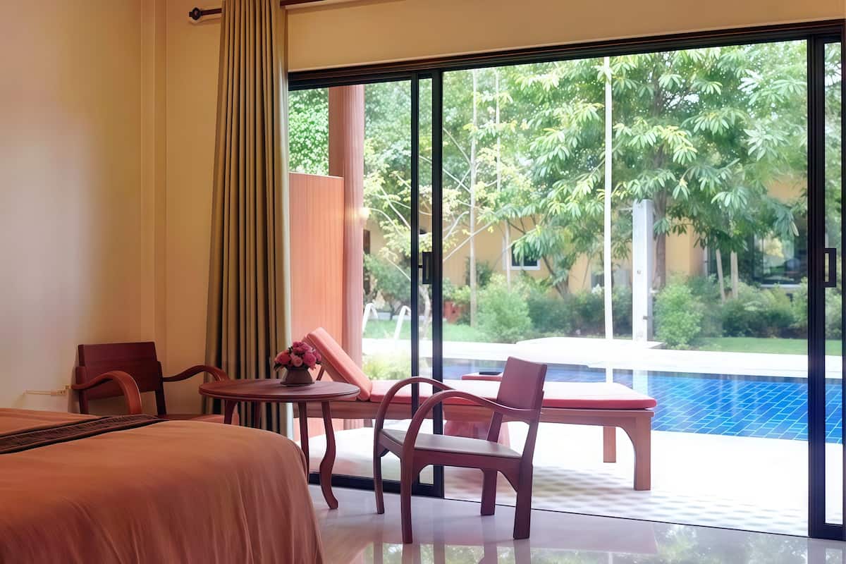 A hotel room with a bed, a table with chairs, a vase of flowers, and a large window with a view of a swimming pool and greenery.