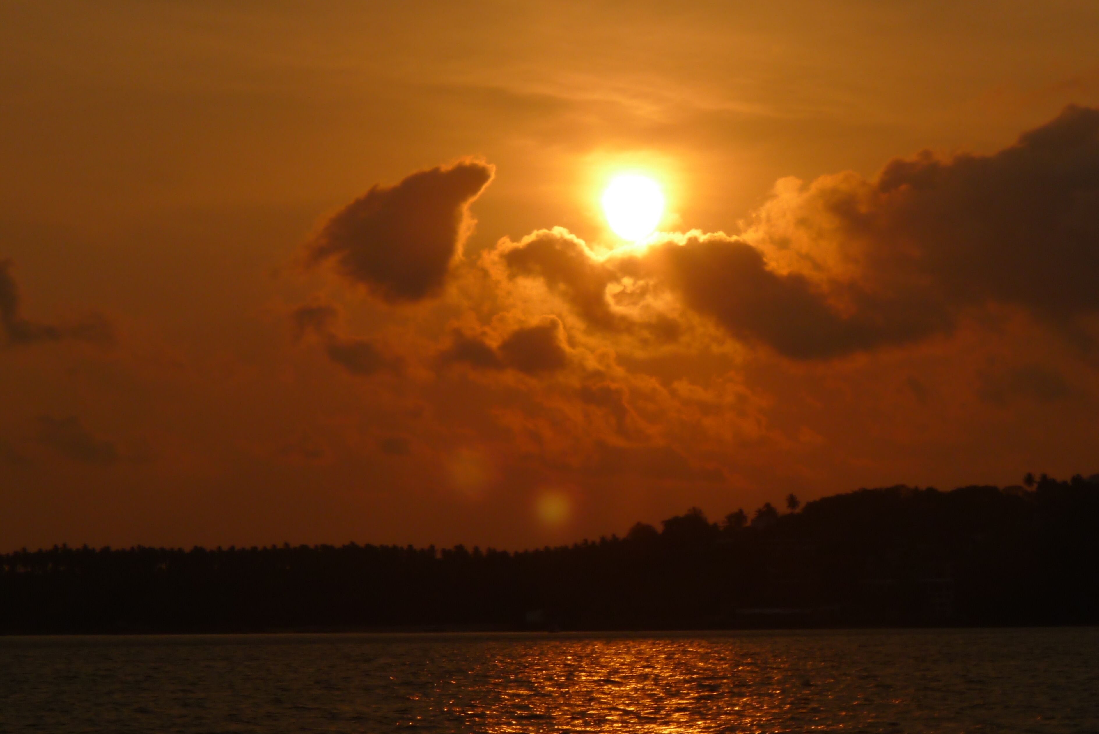 A sunset over a body of water with clouds and a silhouette of a forested area in the background.
