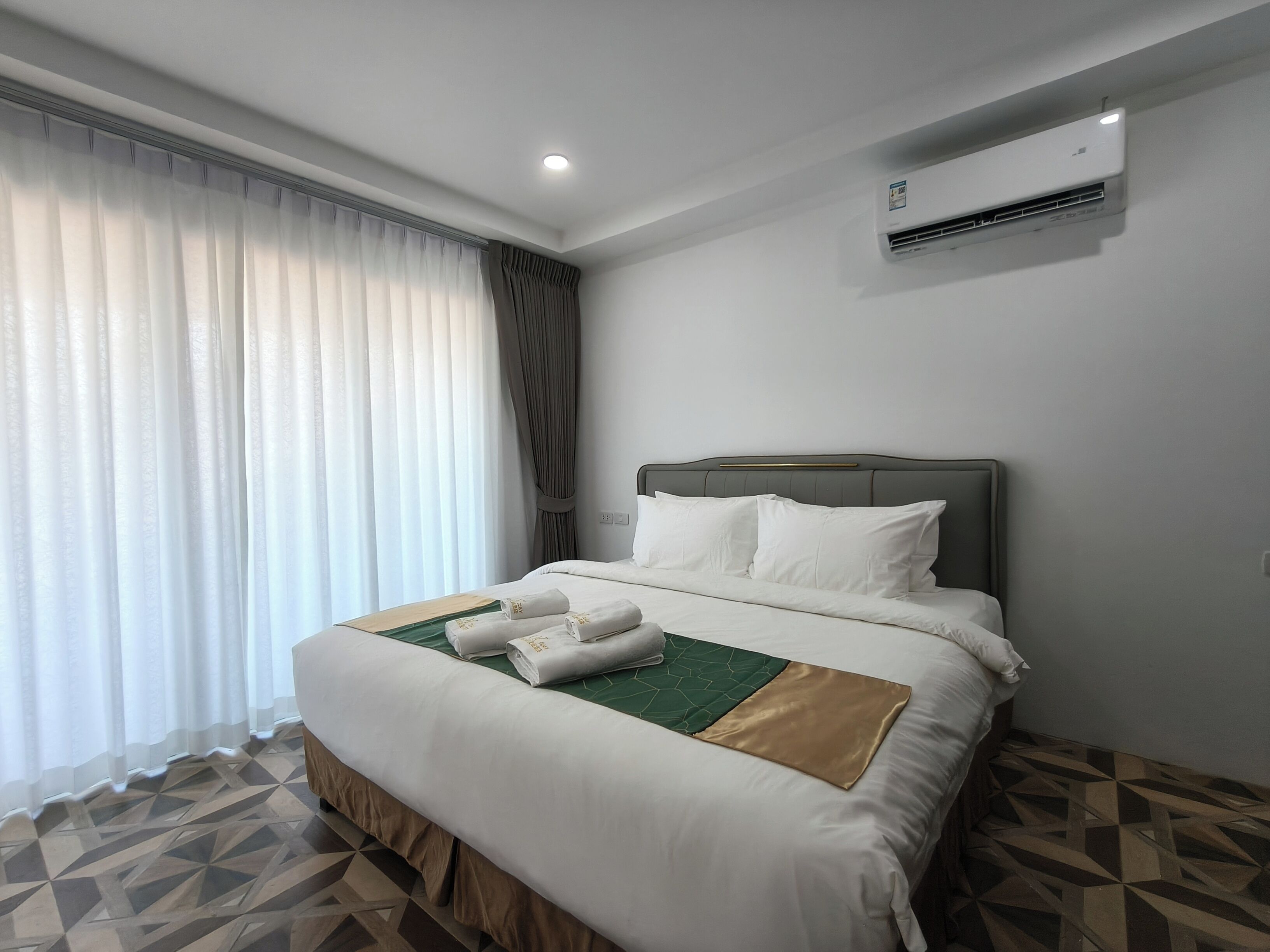 A hotel room with a neatly made bed, a large window with curtains, an air conditioning unit on the wall, and a geometric patterned floor.