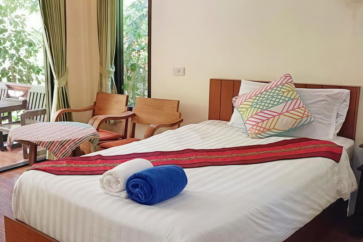 A hotel room with a bed, two chairs, a table, and a view of greenery through the window.