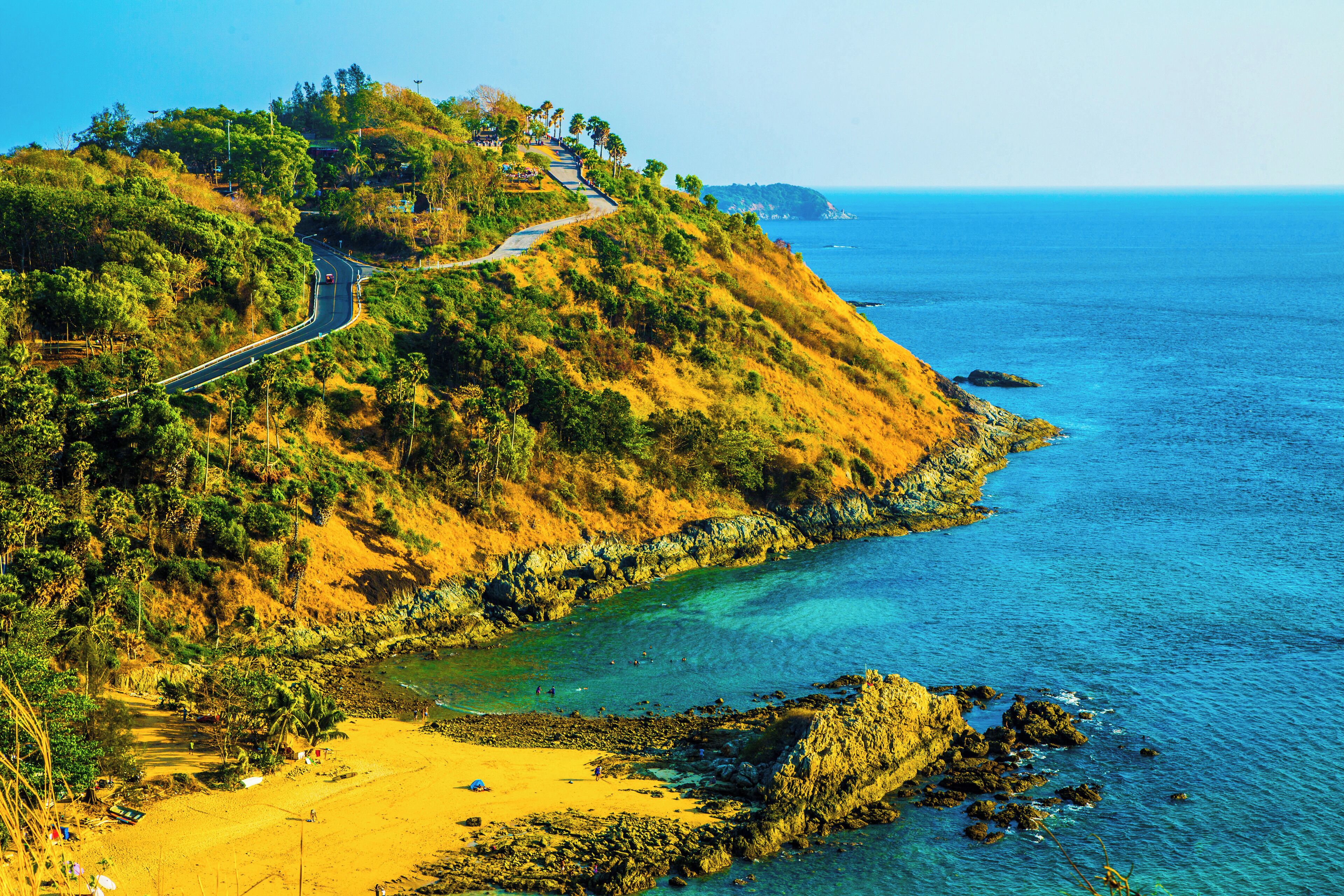 Uma estrada costeira serpenteia por uma paisagem acidentada, com uma praia e litoral rochoso.