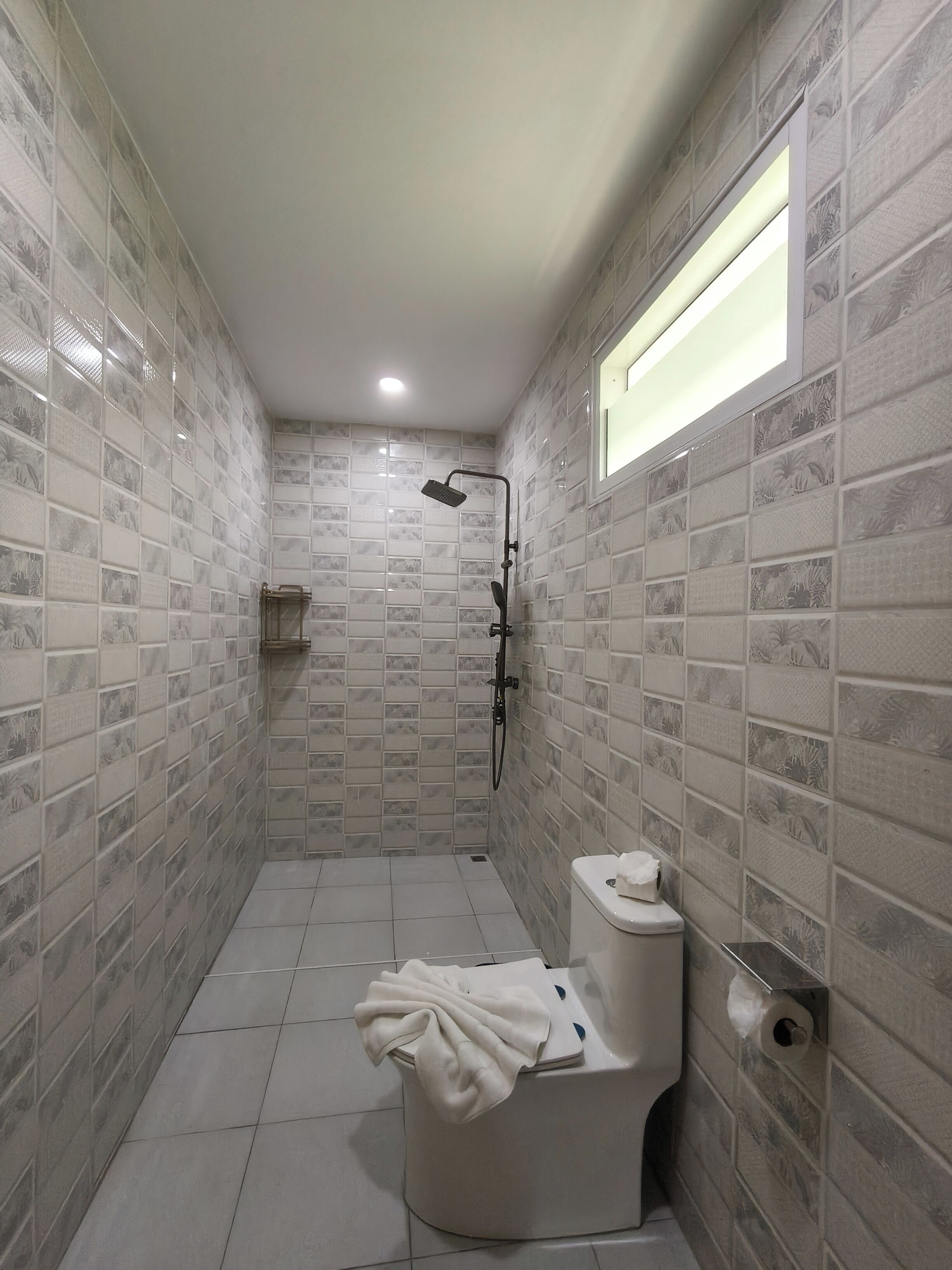 A small, narrow bathroom with tiled walls and floor, a toilet, a shower with a handheld showerhead, and a window providing natural light.