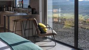 A modern kitchen with a large window, a bar stool, and a chair with a cushion.