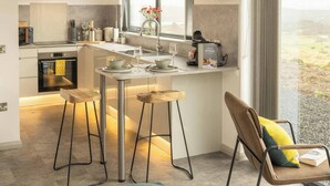 A modern kitchen with a central island, bar stools, a dining table, and a view of the countryside.
