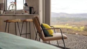 A modern kitchen with a bar area, a chair with a cushion, and a view of the countryside through large windows.