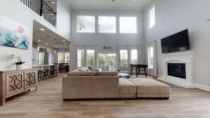 A spacious living room with a large sectional sofa, a fireplace, a flat-screen TV, and a balcony with outdoor seating. - Waterfront Retreat Getaway with Gameroom Firepit Fishing Kayaks (Dickinson)
