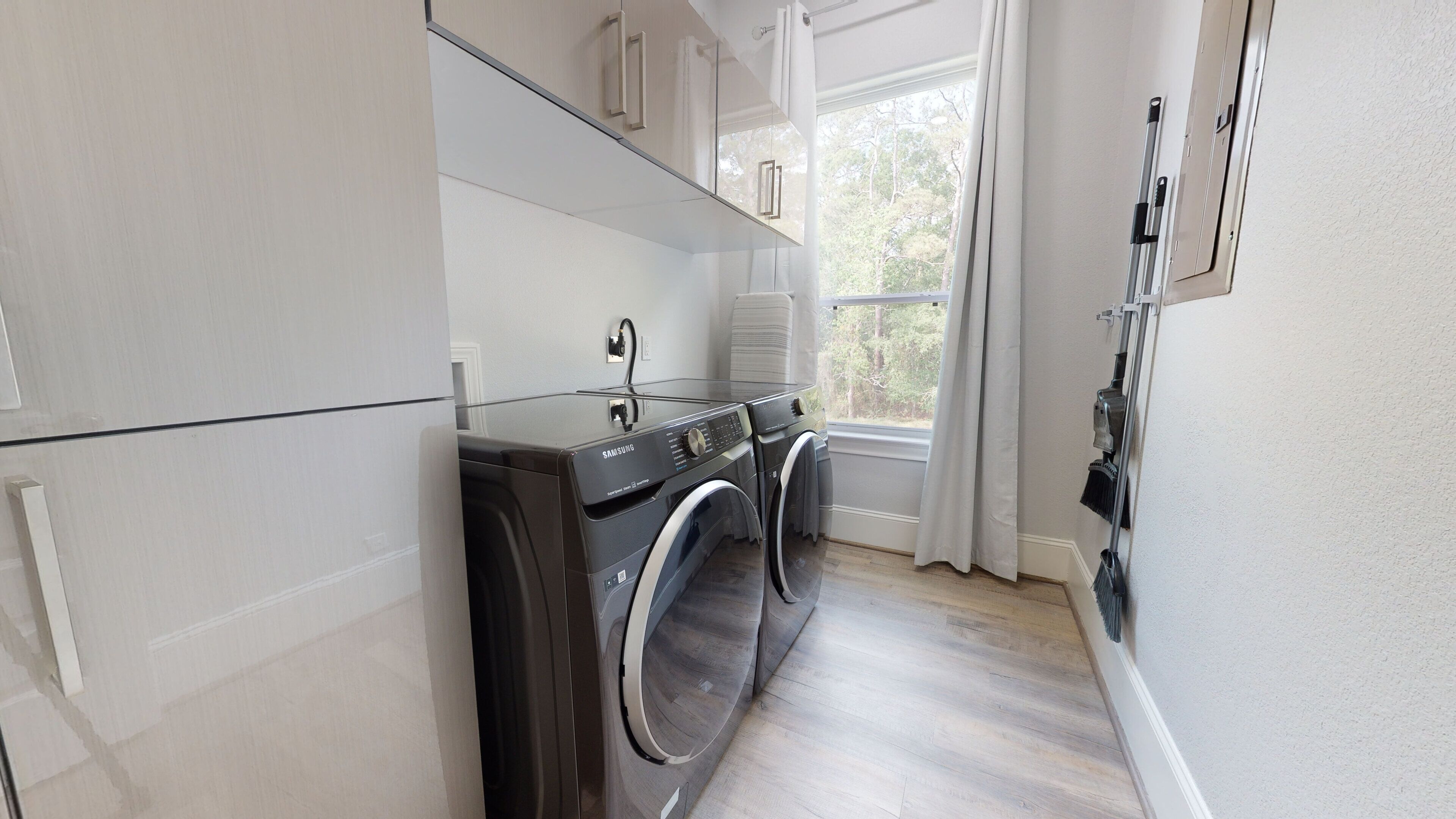 A modern laundry room with two front-loading washing machines, a drying rack, and a window with curtains.