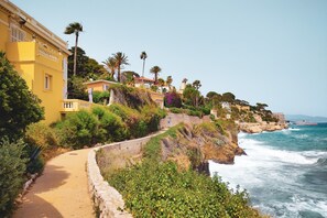 A coastal path with a stone wall, lush greenery, and buildings with balconies overlooking the sea.