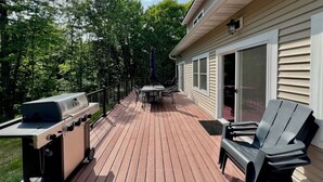 Een houten terras met een barbecue, een tafel en stoelen, omringd door bomen.
