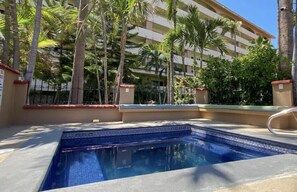 A swimming pool with a tiled edge, surrounded by palm trees and a multi-story building in the background.