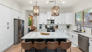 A modern kitchen with white cabinets, stainless steel appliances, a central island with bar stools, and pendant lighting. - KC Oasis POOL W/ HOT Tub Family fun for ALL (Gilbert)