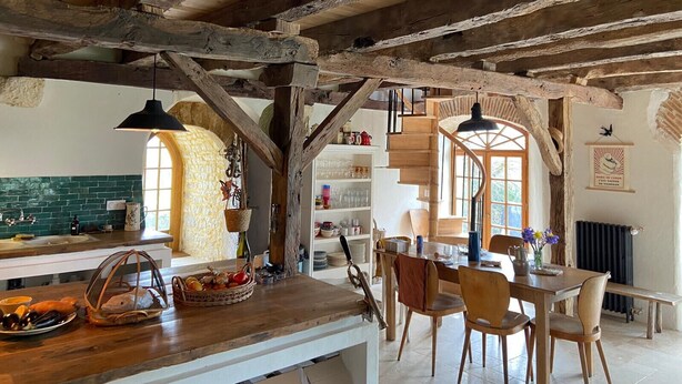 A rustic kitchen with exposed wooden beams, a wooden dining table with chairs, a hanging light fixture, and a window with a view of greenery.